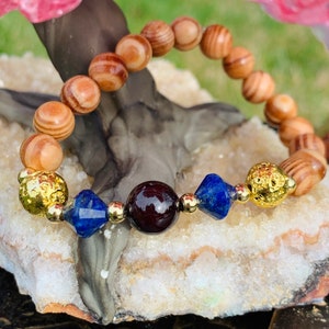 May include: A wooden beaded bracelet with gold, blue, and red gemstone accents. The bracelet is on a white and gray rock surface.