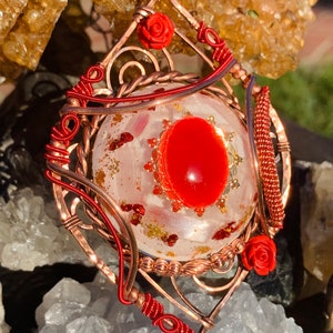 May include: A copper wire wrapped pendant with a red cabochon gemstone and red rose accents. The pendant is set against a white background with gold flecks.