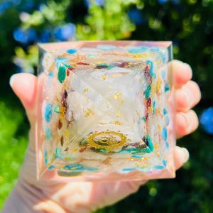 K&ouml;nnte beinhalten: Eine klare Kristallpyramide mit Goldflocken und verschiedenen farbigen Kristallen, die darin eingebettet sind. Die Pyramide hat ein Symbol in der Mitte.