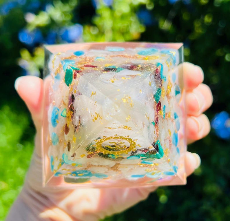 K&ouml;nnte beinhalten: Eine klare Kristallpyramide mit Goldflocken und verschiedenen farbigen Kristallen, die darin eingebettet sind. Die Pyramide hat ein Symbol in der Mitte.