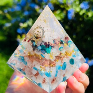 K&ouml;nnte beinhalten: Eine klare Kristallpyramide mit einem goldenen Om-Symbol und verschiedenen Kristallen, die darin eingebettet sind, darunter blaue, gr&uuml;ne und kupferfarbene St&uuml;cke.