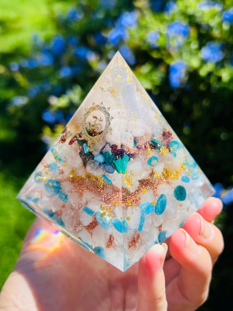 K&ouml;nnte beinhalten: Eine klare Kristallpyramide mit einem goldenen Om-Symbol und verschiedenen Kristallen, die darin eingebettet sind, darunter blaue, gr&uuml;ne und kupferfarbene St&uuml;cke.