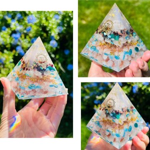 K&ouml;nnte beinhalten: Eine klare Kristallpyramide mit einer Vielzahl von Kristallen und Goldflocken, die darin eingebettet sind. Die Pyramide ist etwa 7,5 cm hoch und steht auf einer Hand.
