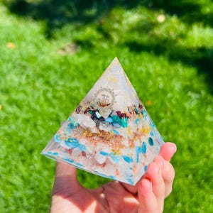 K&ouml;nnte beinhalten: Eine klare pyramidenf&ouml;rmige Kristall mit eingebetteten Steinen, darunter T&uuml;rkis, Rosenquarz und Goldflocken. Die Pyramide wird in einer Hand gehalten.