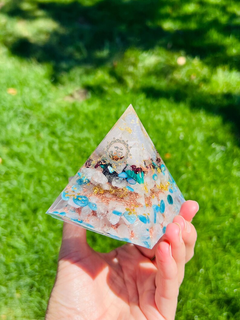 K&ouml;nnte beinhalten: Eine klare pyramidenf&ouml;rmige Kristall mit eingebetteten Steinen, darunter T&uuml;rkis, Rosenquarz und Goldflocken. Die Pyramide wird in einer Hand gehalten.