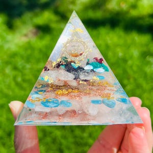 K&ouml;nnte beinhalten: Eine klare Glaspyramide gef&uuml;llt mit verschiedenen Kristallen, darunter Rosenquarz, T&uuml;rkis und Goldflocken. Die Pyramide hat ein goldenes Om-Symbol an der Spitze.