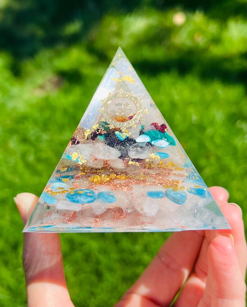 K&ouml;nnte beinhalten: Eine klare Glaspyramide gef&uuml;llt mit verschiedenen Kristallen, darunter Rosenquarz, T&uuml;rkis und Goldflocken. Die Pyramide hat ein goldenes Om-Symbol an der Spitze.