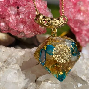 May include: Gold and turquoise resin pendant necklace with a gold Om symbol and a gold chain. The pendant is shaped like a diamond and has a gold floral design on the bail.