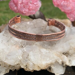 Op de afbeelding: Een koperen draad armband met een gevlochten ontwerp. De armband ligt op een bed van witte kristallen.