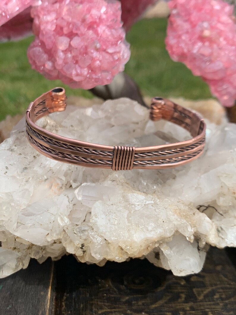 Op de afbeelding: Een koperen draad armband met een gevlochten ontwerp. De armband ligt op een bed van witte kristallen.