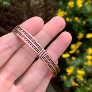 Op de afbeelding: Een koperen armband met een gevlochten ontwerp. De armband is gemaakt van meerdere koperdraden die samen zijn geweven om een touwachtig effect te cre&euml;ren.
