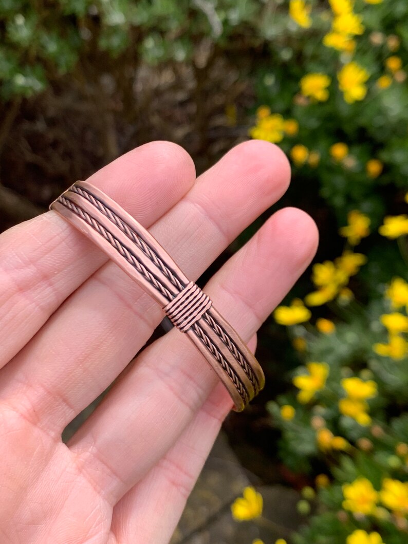 Op de afbeelding: Een koperen armband met een gevlochten ontwerp. De armband is gemaakt van meerdere koperdraden die samen zijn geweven om een touwachtig effect te cre&euml;ren.