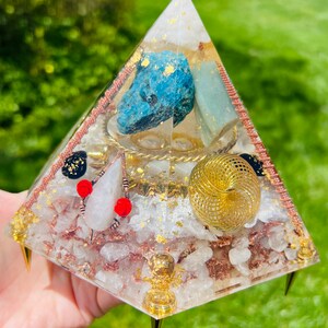 May include: A clear pyramid with a gold wire spiral, a white crystal, a blue crystal, and gold leaf accents. The pyramid is sitting on a hand and has gold metal points on the bottom.