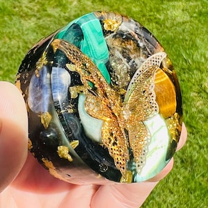 May include: A round, decorative resin piece with a gold filigree butterfly overlay. The resin contains various gemstones in shades of green, blue, and gold, with gold flakes throughout. The piece is held in a hand against a green background.
