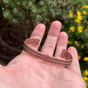 Op de afbeelding: Een koperen armband met een gevlochten ontwerp. De armband is open en heeft een eenvoudige sluiting.