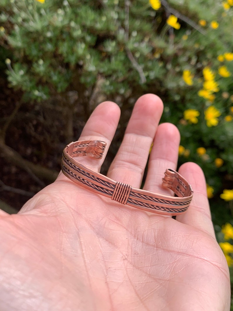 Op de afbeelding: Een koperen armband met een gevlochten ontwerp. De armband is open en heeft een eenvoudige sluiting.