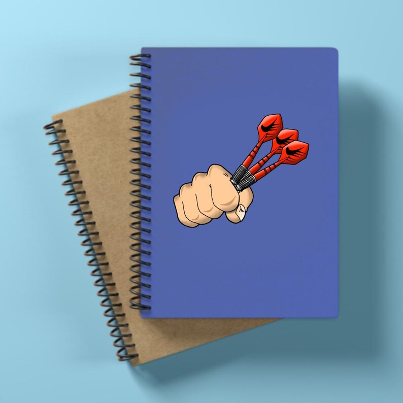 May include: A blue spiral-bound notebook with a cartoon illustration of a fist holding three red darts. A second, brown notebook is partially visible beneath the blue one. The background is a light blue.