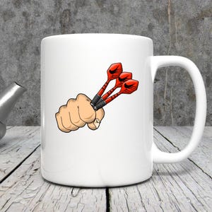 May include: White ceramic mug featuring a cartoon illustration of a hand holding three red and black darts. The darts have a black grip and red flights. The mug has a white handle and is on a wooden surface.