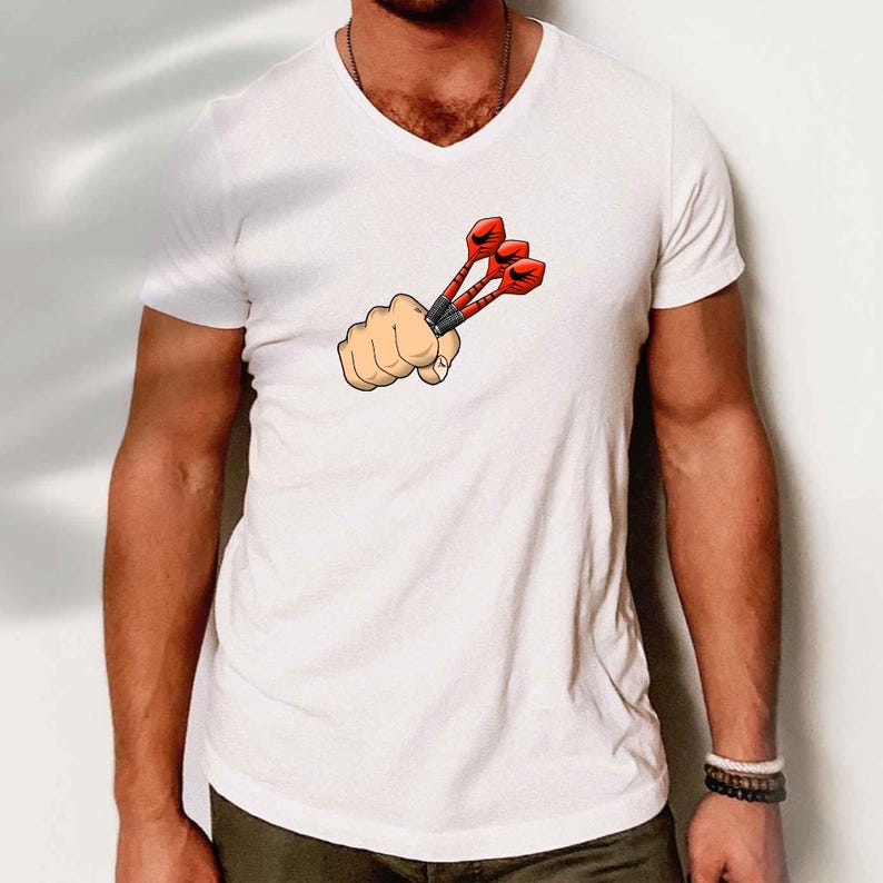 May include: White V-neck t-shirt featuring a cartoon graphic of a clenched fist holding three red and black darts. The darts have red flights and black shafts. The shirt is a casual style.