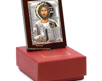 May include: A silver and gold icon of Jesus Christ holding a book, framed in a dark wood frame and displayed in a red box. The icon is labeled "Andcross Orthodox Art Corporation".