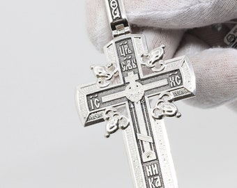 May include: A silver Orthodox cross pendant with intricate engravings and Cyrillic text. The cross features a detailed design with floral accents and a hinged bail for attaching to a chain. The pendant is held against a white background.