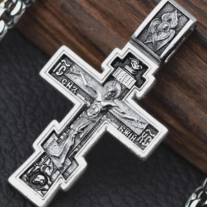 Könnte beinhalten: Ein detaillierter silberner Anhänger in Form eines Kreuzes mit einer eingravierten Darstellung von Jesus Christus. Das Kreuz hat komplizierte Designs und religiöse Ikonografie, mit einer Kette im Hintergrund sichtbar. Der Anhänger ist wahrscheinlich aus einer Metalllegierung.