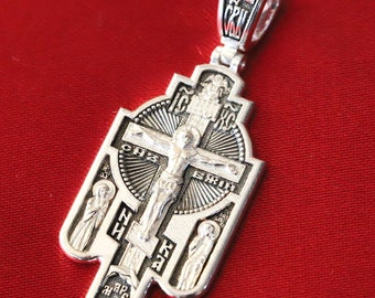 May include: A silver pendant in the shape of a cross with intricate details. The cross features a depiction of Jesus Christ on the crucifix, surrounded by Cyrillic lettering. The pendant has a loop at the top for attaching to a chain.