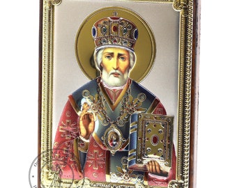 May include: A silver and gold icon of Saint Nicholas, also known as Santa Claus, with a red and blue robe, a gold crown, and a book in his hand. The icon is framed in a wooden frame with a gold trim.