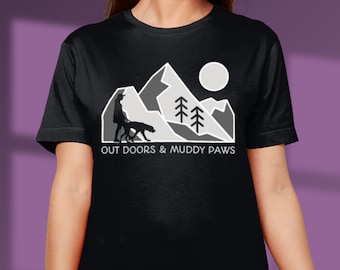 May include: Black t-shirt with a white graphic of a person walking a dog in front of a mountain range with a sun and trees. The text "OUT DOORS & MUDDY PAWS" is below the graphic.