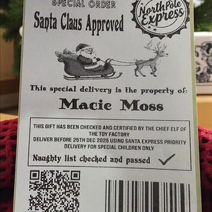 May include: A Christmas delivery tag from the Lapland Toy Factory, approved by Santa Claus. The tag is addressed to Macie Moss and includes a QR code and barcode. The tag is placed on a red knitted surface.