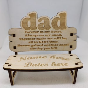 May include: A light-coloured wooden bench-shaped memorial with the word "dad" at the top. The bench has engraved text: "Forever in my heart, Always on my mind..." and space for a name and dates. The bench is made of light-coloured wood.