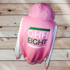 Op de afbeelding: Roze en witte baseballjas met een groen en wit glitterontwerp met de tekst "OHH EIGHT" op de rug.