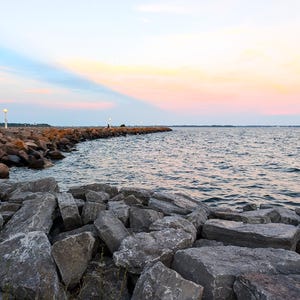 May include: A coastal scene with a stone breakwater stretching into the sea. The sky shows a gradient of colours, from blue to pink and orange. The foreground is filled with large grey rocks, with a calm sea in the background.