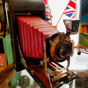May include: Vintage black and red folding camera with brass accents. The camera features a red bellows, a brass lens, and a viewfinder. The camera is open, revealing the lens and internal components. The camera is displayed on a glass surface.
