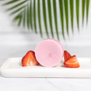 Puede incluir: Una barra de jabón redonda de color rosa sobre un plato blanco con dos rodajas de fresas rojas.