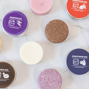 Pode incluir: Uma vista superior de várias barras de champô e condicionador. As barras são redondas e vêm em diferentes cores e texturas. Algumas estão em pequenas latas redondas de metal com rótulos que dizem "Shampoo Bar" e "Conditioner Bar". A marca "bar bara" é visível.