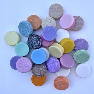 Puede incluir: Una colección de pastillas de jabón redondas y coloridas en varios tonos de azul, verde, rosa, morado y marrón. Las pastillas de jabón están dispuestas en un patrón circular sobre una superficie blanca.
