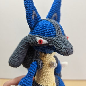 May include: A crocheted plush toy of a blue and grey Pokemon character with red eyes. The toy is made with yarn and has a yellow chest.