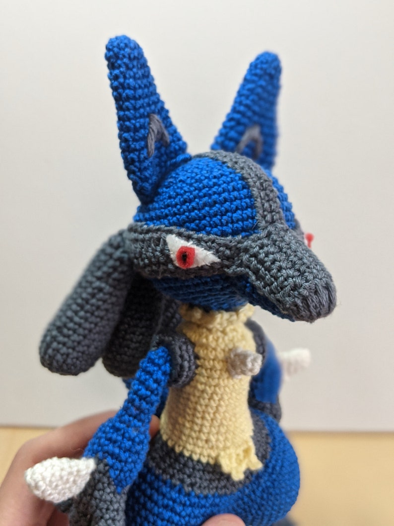 May include: A crocheted plush toy of a blue and grey Pokemon character with red eyes. The toy is made with yarn and has a yellow chest.