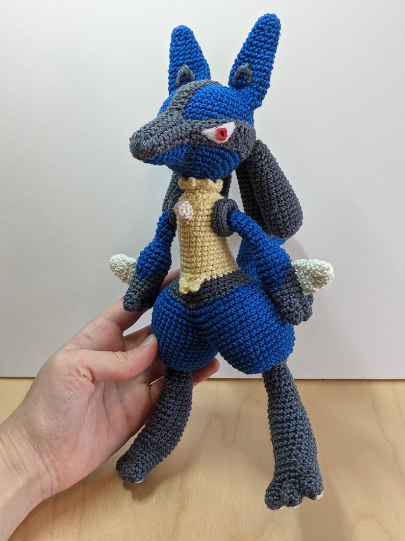 May include: A blue and grey crocheted plush toy resembling a Pokemon character. The toy has a yellow chest and red eyes.