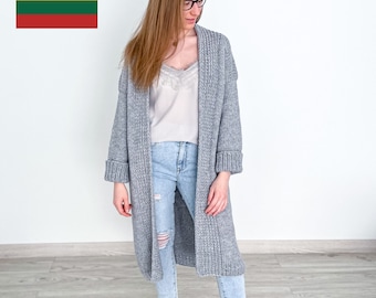 May include: A woman wearing a long, gray knitted cardigan, light blue jeans, and black sneakers. She is standing in front of a white wall.