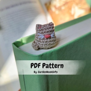 May include: A crochet cat bookmark in a light brown color with a pink nose and blue eyes. The bookmark is sitting on top of a green book with the text "PDF Pattern By: GardenNookGifts".