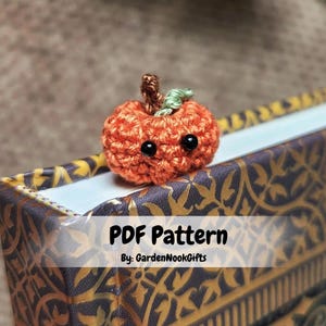May include: A crocheted pumpkin with a green stem and black eyes. The pumpkin is sitting on a book with the text "PDF Pattern By: GardenNookGifts".