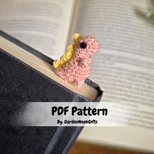 Puede incluir: Marcapáginas de dinosaurio tejido a ganchillo en rosa y amarillo, colocado en un libro. La imagen incluye el texto "PDF Pattern" y "By: GardenNookGifts". Un accesorio encantador para los amantes de los libros.
