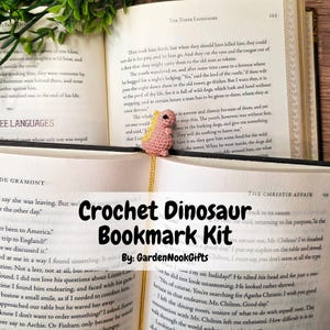 May include: A small, pink, crocheted dinosaur bookmark with yellow spikes along its back, attached to a thin yellow cord. The bookmark is placed between the pages of an open book, with the dinosaur head visible above the top edge of the pages.