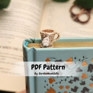 Op de afbeelding: Een gehaakt theekopje-bladwijzer met een theezakje dat aan de zijkant hangt. De bladwijzer ligt op een blauw boek met een bloemenpatroon. De tekst "PDF Pattern" en "By: GardenNookGifts" wordt onder de bladwijzer weergegeven.
