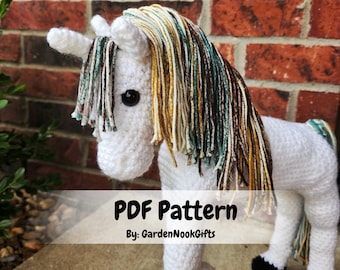 May include: Crochet pattern for a white unicorn with a long, multi-colored mane. The mane is made of teal, brown, and yellow yarn. The pattern is available as a PDF download. "PDF Pattern By: GardenNookGifts"