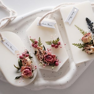 May include: Three white wax melts with dried flowers and greenery embedded in the wax. Each melt has a small tag with the text "Anella Flowers" written on it.