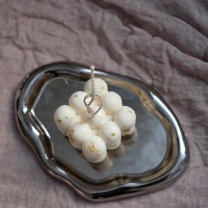 May include: A white candle with gold flecks, shaped like a cluster of spheres, sits on a silver tray. The candle has a white string wick.