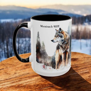 May include: White ceramic mug with a black handle and rim. The mug features a watercolor illustration of a wolf in a snowy forest setting. The text "Matriarch 907F" is printed on the mug.
